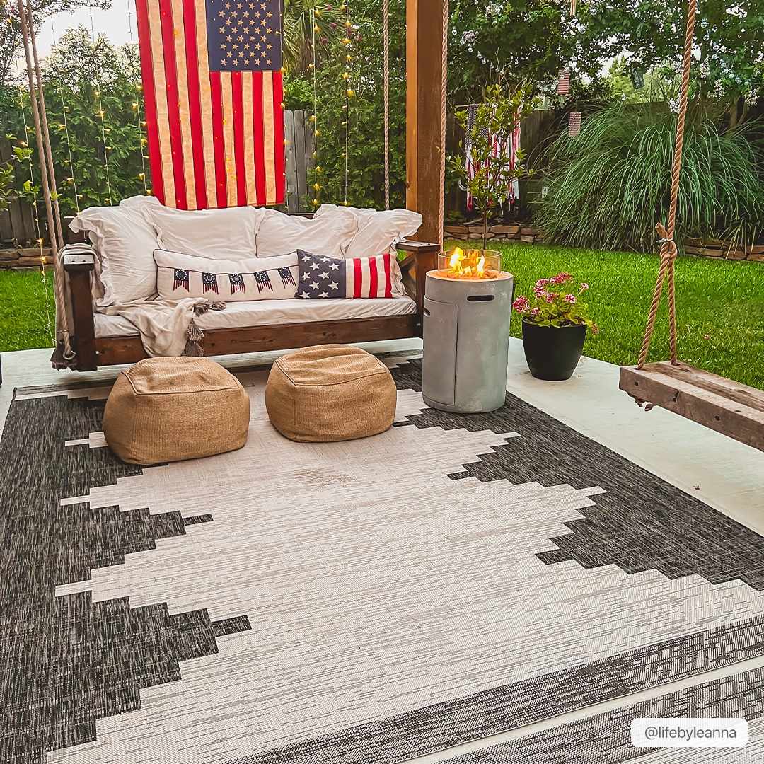 Outdoor patio with a wooden swing, cushions, and a rug on a grassy area.